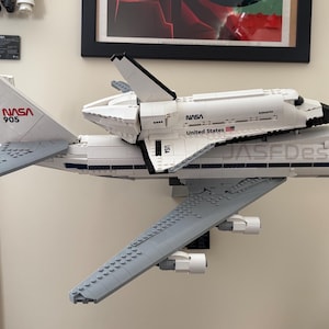 May include: A white LEGO model of a NASA space shuttle atop a Boeing 747 aircraft. The model features the NASA logo and the text "United States". The aircraft has the text "JASEDesign" on the side.