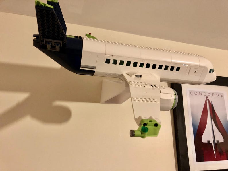 May include: A white and blue Lego airplane model mounted on a white wall shelf. The airplane has a green tail and is partially obscured by a shadow. A framed poster with the text "CONCORDE" is visible in the background.
