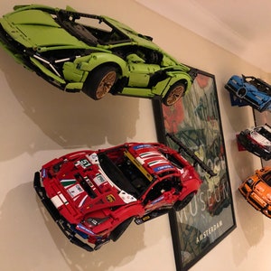 May include: Five different Lego car models are displayed on a wall. The cars are a green Lamborghini, a red Ferrari, a blue Bugatti, a white Porsche, and an orange Porsche. The cars are all mounted on the wall with a black bracket.