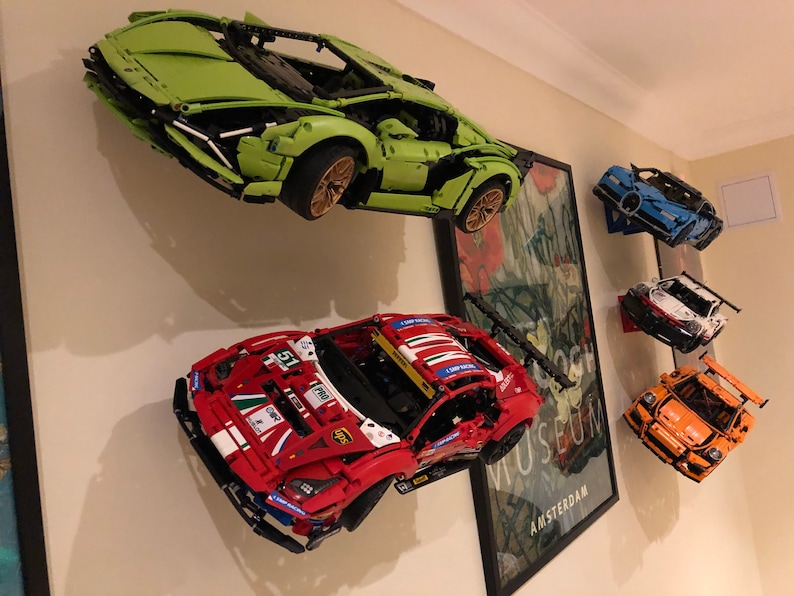 May include: Five different Lego car models are displayed on a wall. The cars are a green Lamborghini, a red Ferrari, a blue Bugatti, a white Porsche, and an orange Porsche. The cars are all mounted on the wall with a black bracket.