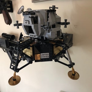 May include: A silver and black Lego model of the Apollo Lunar Module, with a gold-colored base and the words "UNITED STATES" printed on it. The model is mounted on a wall.