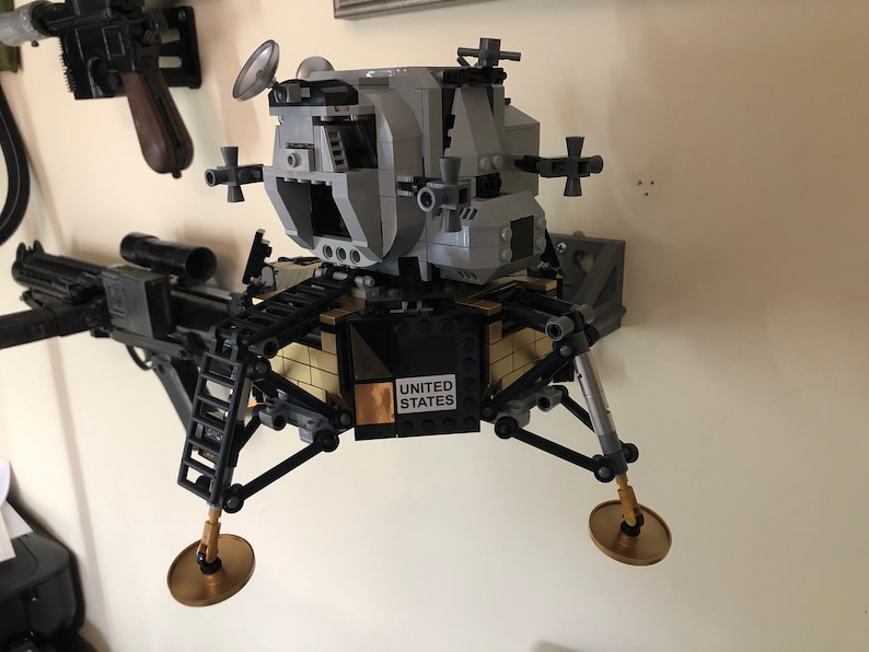 May include: A silver and black Lego model of the Apollo Lunar Module, with a gold-colored base and the words "UNITED STATES" printed on it. The model is mounted on a wall.