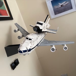 May include: A white and blue toy space shuttle model mounted on top of a gray airplane model. The toy has black accents and is displayed against a neutral wall. The model is a detailed replica of a space shuttle.