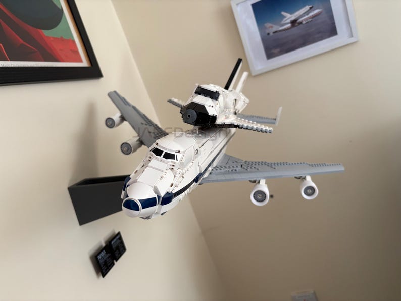 May include: A white and blue toy space shuttle model mounted on top of a gray airplane model. The toy has black accents and is displayed against a neutral wall. The model is a detailed replica of a space shuttle.