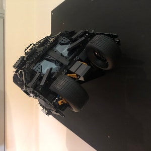 May include: A black Lego model of the Batmobile from the Batman movie series. The model is mounted on a black wall and is facing the camera. The Batmobile has a detailed design with black and gold accents.