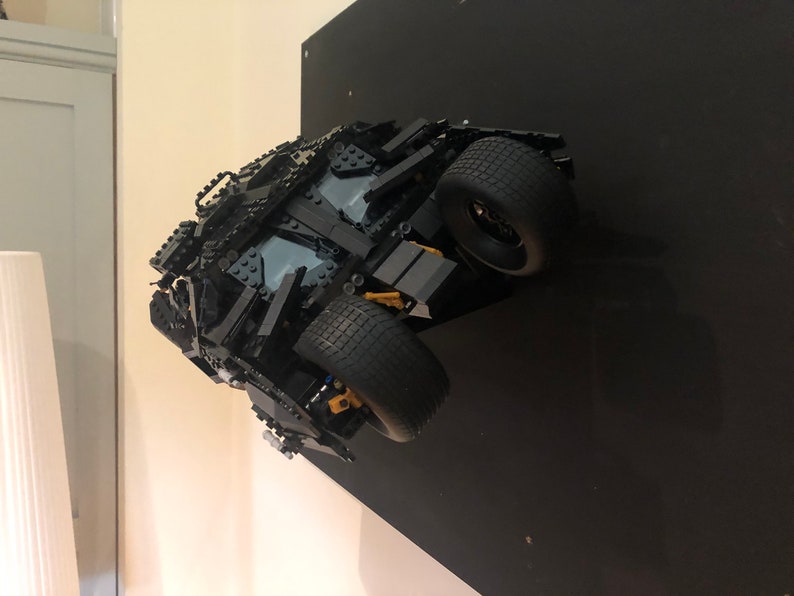 May include: A black Lego model of the Batmobile from the Batman movie series. The model is mounted on a black wall and is facing the camera. The Batmobile has a detailed design with black and gold accents.