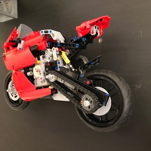 Op de afbeelding: Een rood en zwart Lego-motorfietsmodel met een gedetailleerde motor en kettingaandrijving. De motorfiets hangt in de lucht tegen een zwarte achtergrond.