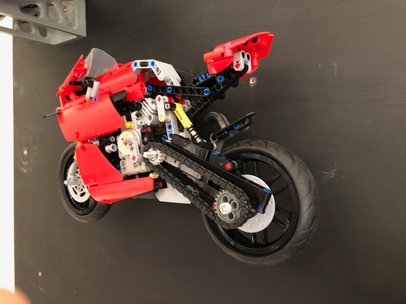 Op de afbeelding: Een rood en zwart Lego-motorfietsmodel met een gedetailleerde motor en kettingaandrijving. De motorfiets hangt in de lucht tegen een zwarte achtergrond.