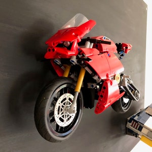 Op de afbeelding: Een rood Lego-motorfietsmodel met een zwarte voorband en een kleinere zwarte achterband. De motorfiets is gemonteerd op een zwarte muur.