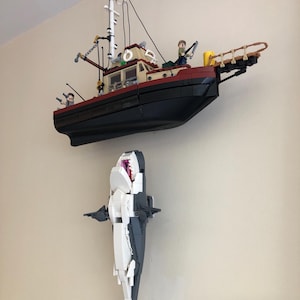 May include: A Lego model of a fishing boat with a black hull, tan cabin, and white mast, mounted above a Lego shark with an open mouth. The shark is white and gray.