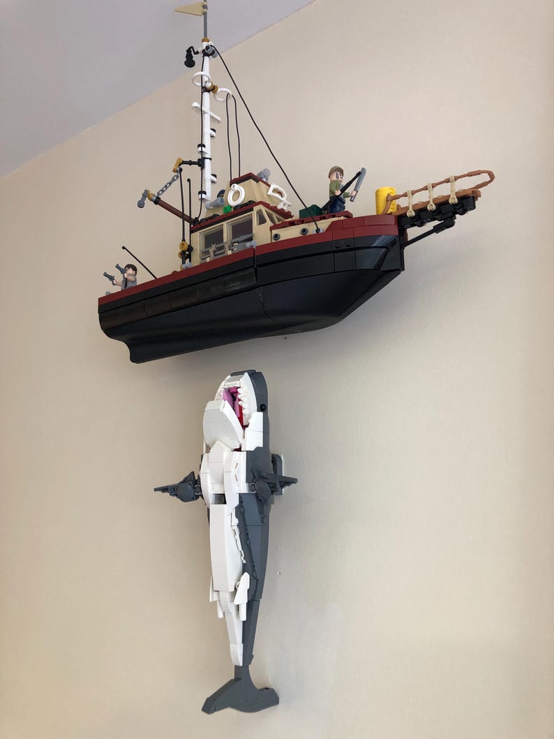 May include: A Lego model of a fishing boat with a black hull, tan cabin, and white mast, mounted above a Lego shark with an open mouth. The shark is white and gray.