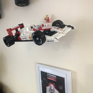 Op de afbeelding: Een wit en rode Lego raceauto met zwarte wielen en het woord "McLaren" op de zijkant. Een klein Lego figuur staat bovenop de auto. Een ingelijste foto van een raceauto staat eronder.