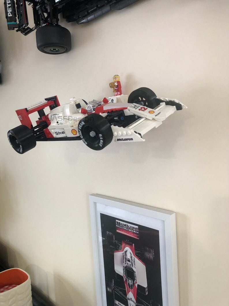 Op de afbeelding: Een wit en rode Lego raceauto met zwarte wielen en het woord "McLaren" op de zijkant. Een klein Lego figuur staat bovenop de auto. Een ingelijste foto van een raceauto staat eronder.