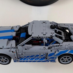 May include: A gray and blue Lego Technic car model with a large rear spoiler. The car has blue stripes and accents, and the wheels are visible. The text "ASF Design" is on the side of the car.