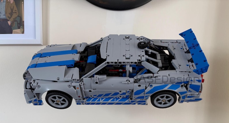May include: A gray and blue Lego Technic car model with a large rear spoiler. The car has blue stripes and accents, and the wheels are visible. The text "ASF Design" is on the side of the car.