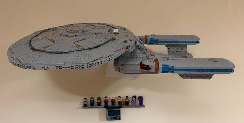 Puede incluir: Un gran modelo Lego gris de la nave espacial Star Trek USS Enterprise. El modelo est&aacute; montado en una pared, con una peque&ntilde;a exhibici&oacute;n de figuras de Lego debajo. La nave presenta una construcci&oacute;n y un dise&ntilde;o detallados.