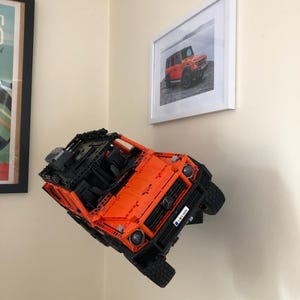 May include: An orange and black Lego model of a Mercedes-Benz G-Wagon is mounted on a wall. The model is suspended in mid-air, as if it is driving up a wall. The model has a black license plate that reads "G 63 AMG".