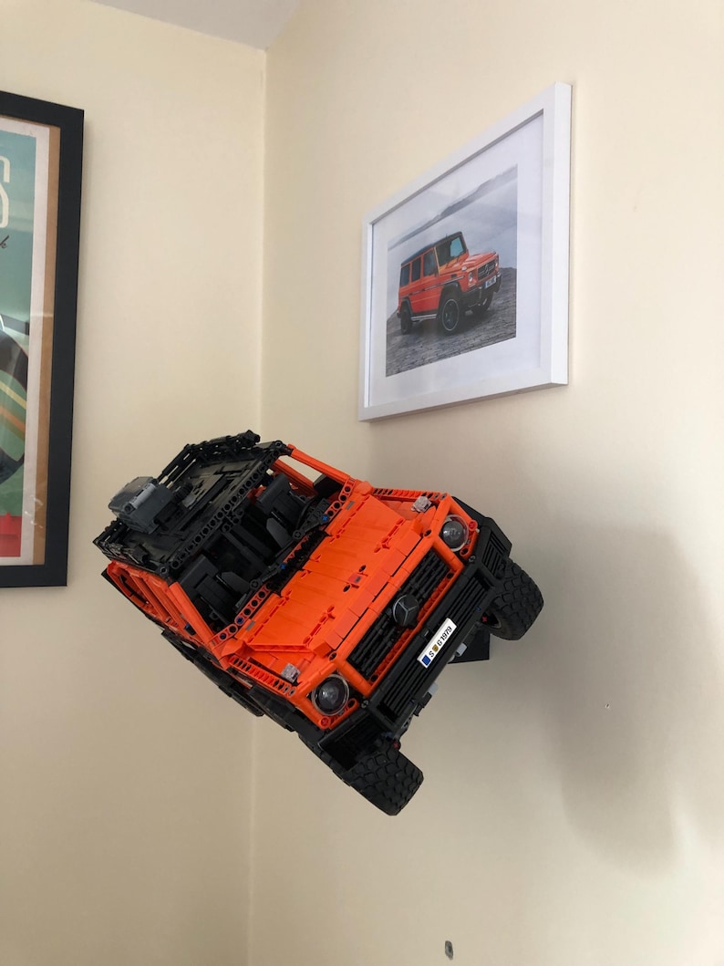 May include: An orange and black Lego model of a Mercedes-Benz G-Wagon is mounted on a wall. The model is suspended in mid-air, as if it is driving up a wall. The model has a black license plate that reads "G 63 AMG".
