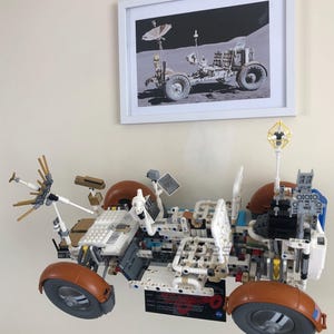 May include: A detailed LEGO model of a lunar rover, featuring brown, white, and gray components. The model has large wheels and various antennas and panels. Above it is a framed black and white photo of a lunar rover. The text "LEGO Not Included" is at the bottom.