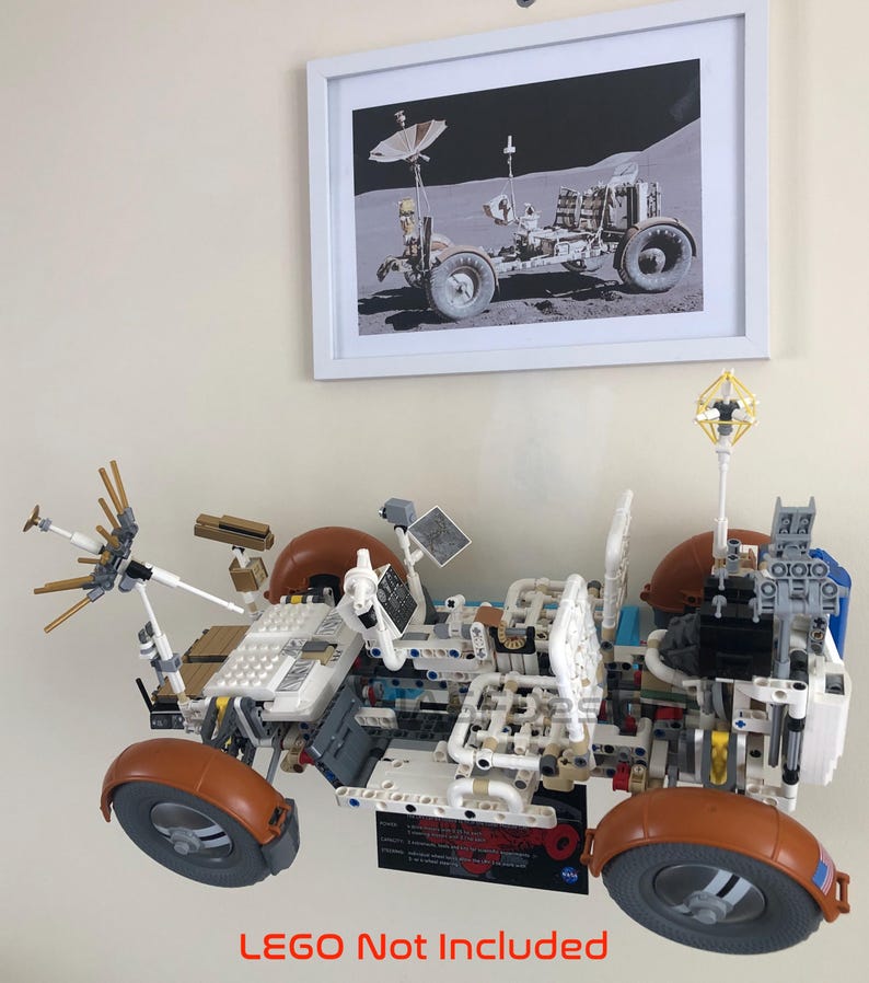 May include: A detailed LEGO model of a lunar rover, featuring brown, white, and gray components. The model has large wheels and various antennas and panels. Above it is a framed black and white photo of a lunar rover. The text "LEGO Not Included" is at the bottom.