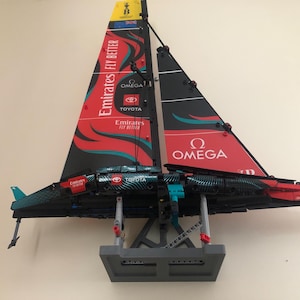 K&ouml;nnte beinhalten: Ein schwarzes und rotes Lego-Modell eines Segelboots mit den Worten "Emirates Fly Better" und "Omega" auf den Segeln. Das Modell ist auf einem grauen St&auml;nder montiert.