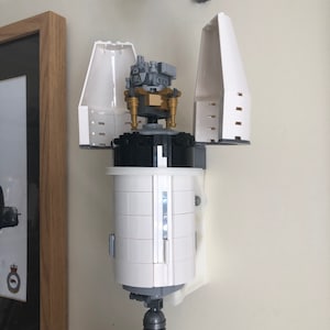 May include: A white Lego model of a rocket ship with a gray and gold Lego lunar lander inside. The rocket ship is mounted on a white wall mount.