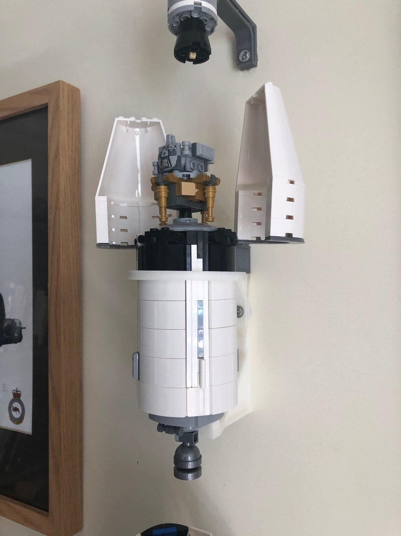 May include: A white Lego model of a rocket ship with a gray and gold Lego lunar lander inside. The rocket ship is mounted on a white wall mount.
