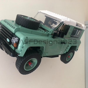 May include: A green Lego Land Rover Defender model with a white roof and black tires. The model has the word "DEFENDER" printed on the hood.