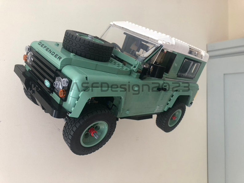 May include: A green Lego Land Rover Defender model with a white roof and black tires. The model has the word "DEFENDER" printed on the hood.