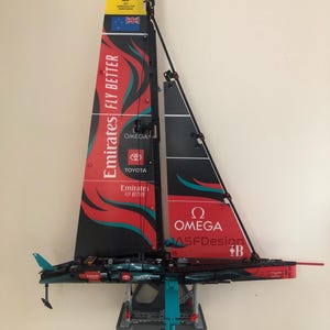 K&ouml;nnte beinhalten: Ein rotes, schwarzes und t&uuml;rkisfarbenes LEGO-Modell eines Segelboots mit den Worten "Emirates Fly Better", "Omega", "Toyota" und "MASFDesign" auf den Segeln. Das Segelboot steht auf einem grauen St&auml;nder.
