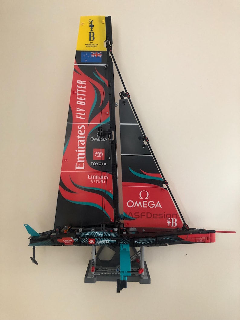 K&ouml;nnte beinhalten: Ein rotes, schwarzes und t&uuml;rkisfarbenes LEGO-Modell eines Segelboots mit den Worten "Emirates Fly Better", "Omega", "Toyota" und "MASFDesign" auf den Segeln. Das Segelboot steht auf einem grauen St&auml;nder.