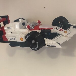 May include: A red, white, and black Lego model of a Formula One race car. The car has the number 12 on the side and the word "McLaren" on the bottom. The car is made of plastic and has a detailed design.