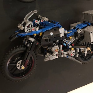 May include: A blue and black Lego motorcycle model with a BMW logo on the side. The motorcycle has two side cases and a top case. The motorcycle is on a black background.