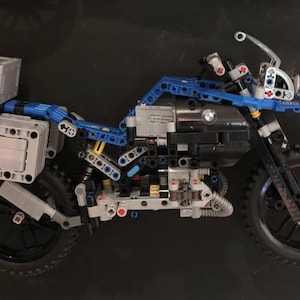 May include: A blue and gray Lego motorcycle model with a detailed engine and luggage racks. The motorcycle has a black seat and black wheels with knobby tires.