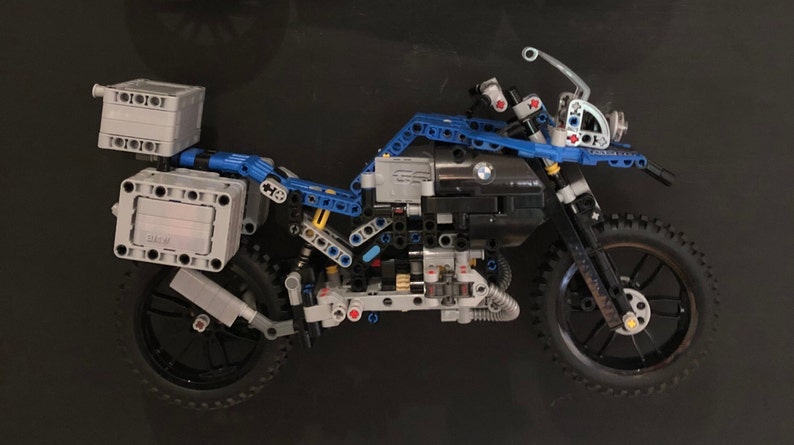 May include: A blue and gray Lego motorcycle model with a detailed engine and luggage racks. The motorcycle has a black seat and black wheels with knobby tires.