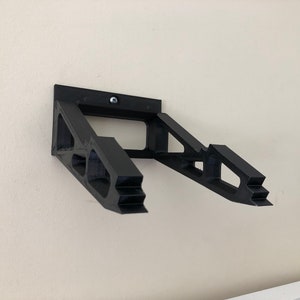 May include: A black 3D printed wall mount for a laptop or tablet. The mount has a simple design with a rectangular base and two angled arms that extend out to support the device.