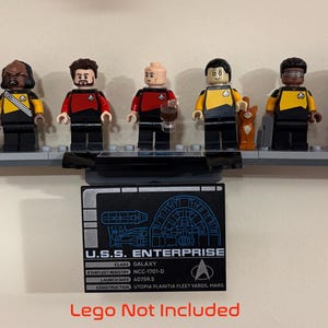 Puede incluir: Una colecci&oacute;n de minifiguras de Lego de Star Trek expuestas sobre una base Lego gris. Las figuras representan a varios personajes con sus uniformes. Una placa negra debajo dice "U.S.S. Enterprise" con detalles t&eacute;cnicos. El texto "Lego Not Included" est&aacute; en la parte inferior.