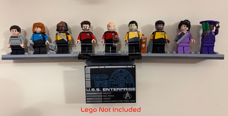 Puede incluir: Una colecci&oacute;n de minifiguras de Lego de Star Trek expuestas sobre una base Lego gris. Las figuras representan a varios personajes con sus uniformes. Una placa negra debajo dice "U.S.S. Enterprise" con detalles t&eacute;cnicos. El texto "Lego Not Included" est&aacute; en la parte inferior.