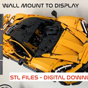 Puede incluir: Un modelo de coche Lego amarillo con detalles negros. El coche est&aacute; montado en una pared con un soporte impreso en 3D verde. La imagen incluye el texto "WALL MOUNT TO DISPLAY" y "STL FILES - DIGITAL DOWNLOAD".