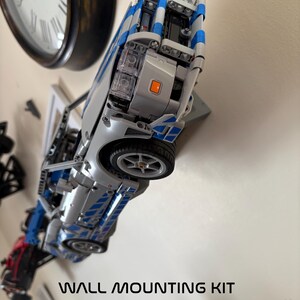May include: A silver and blue toy car mounted on a wall, with the text "WALL MOUNTING KIT" below. The car has visible wheels and detailed features. A clock is visible in the background.