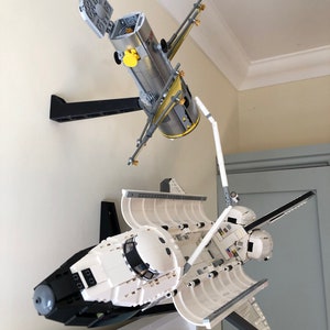 May include: A Lego model of a space shuttle and a satellite, mounted on a wall. The shuttle is white and black, with open cargo doors. The satellite is gray and yellow, with solar panels extended. The models are displayed on black brackets.