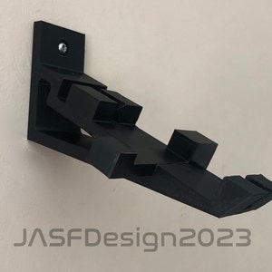 May include: A black 3D printed wall mount designed to hold a video game controller. The mount is attached to the wall with a screw. The mount has a unique design with multiple angles and curves. The text "JASFDesign2023" is visible in the bottom of the image.