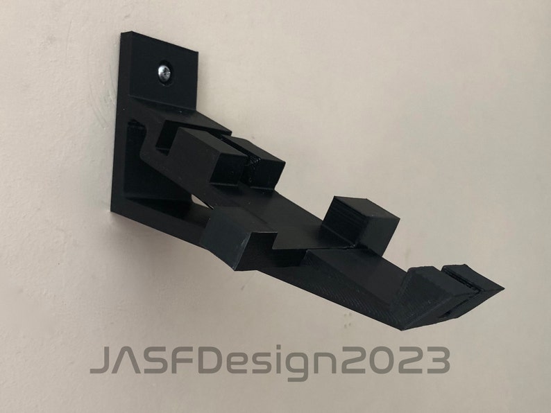 May include: A black 3D printed wall mount designed to hold a video game controller. The mount is attached to the wall with a screw. The mount has a unique design with multiple angles and curves. The text "JASFDesign2023" is visible in the bottom of the image.