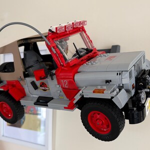 May include: A brick-built toy vehicle model featuring a gray and red exterior with a tan soft top, a curved antenna, and a roof-mounted light bar. The side of the vehicle displays the number 12 and a circular Jurassic Park logo on the door panel.