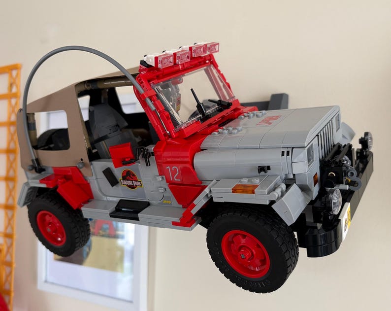 May include: A brick-built toy vehicle model featuring a gray and red exterior with a tan soft top, a curved antenna, and a roof-mounted light bar. The side of the vehicle displays the number 12 and a circular Jurassic Park logo on the door panel.