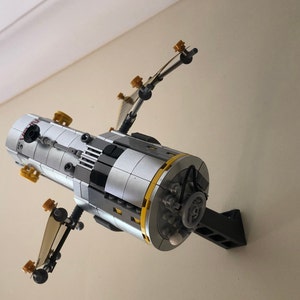 May include: A Lego model of a space station, featuring a cylindrical body with silver, black, and yellow accents. The model has multiple protruding solar panels and is mounted on a black bracket against a beige wall.
