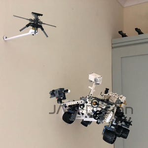 Peut inclure: Un mod&egrave;le de rover martien Lego blanc et gris avec un bras robotique et des roues. Le rover est mont&eacute; sur un mur blanc avec un mod&egrave;le d'h&eacute;licopt&egrave;re Lego blanc et gris mont&eacute; sur le mur au-dessus.
