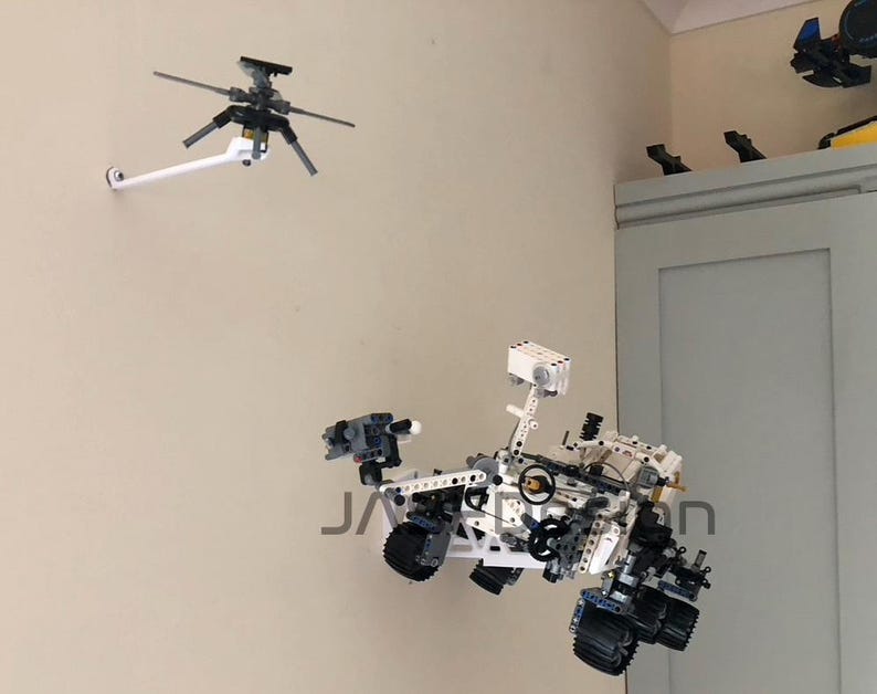 Peut inclure: Un mod&egrave;le de rover martien Lego blanc et gris avec un bras robotique et des roues. Le rover est mont&eacute; sur un mur blanc avec un mod&egrave;le d'h&eacute;licopt&egrave;re Lego blanc et gris mont&eacute; sur le mur au-dessus.