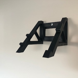 Op de afbeelding: Zwarte wandbeugel voor een videogameconsole. De beugel is gemaakt van plastic en heeft een driehoekige vorm. Hij is ontworpen om de console stevig aan de muur te bevestigen.
