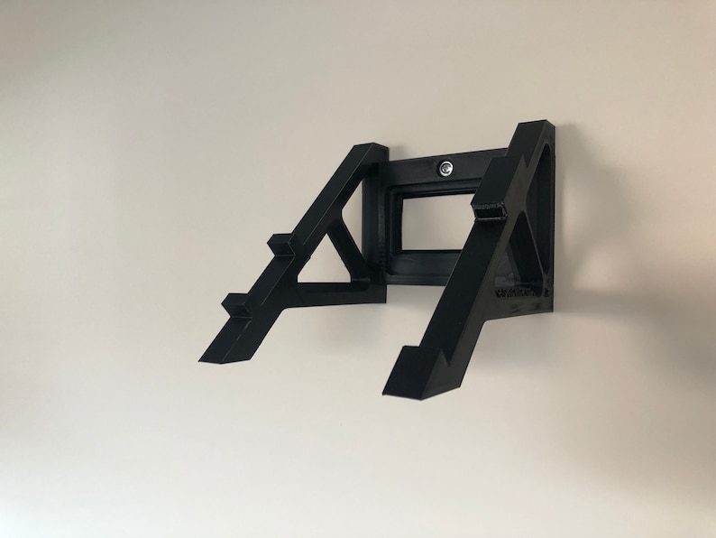Op de afbeelding: Zwarte wandbeugel voor een videogameconsole. De beugel is gemaakt van plastic en heeft een driehoekige vorm. Hij is ontworpen om de console stevig aan de muur te bevestigen.